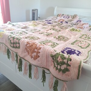 Può includere: Una coperta rosa all'uncinetto fatta a mano con motivi quadrati nei toni del rosa, verde e marrone. La coperta ha un bordo smerlato con nappe rosa e verdi. La coperta è drappeggiata su un telaio del letto bianco.