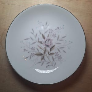 May include: A white ceramic plate with a silver rim features a delicate floral design. The design includes a pink rose with gray leaves and small white and blue flowers.