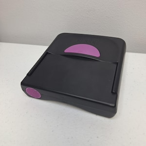 Peut inclure: Appareil électronique en plastique noir avec un accent violet. L'appareil a une forme rectangulaire avec un bouton violet incurvé et un petit bouton violet sur le côté. Le mot "STODD" est imprimé en haut.