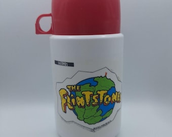 Puede incluir: Termo blanco con tapa y asa rojas. El termo presenta un gráfico colorido del logotipo de dibujos animados de los Picapiedra con un diseño de mapa del mundo verde y azul. La palabra "THERMOS" está impresa en el lateral.