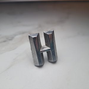 May include: A silver metal letter "H" with a geometric design. The letter is standing upright and has a slightly textured surface. The background is blurred, focusing attention on the letter.