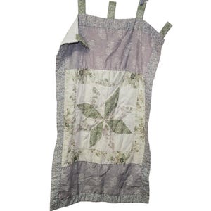 May include: A quilted, floral-patterned wall hanging in shades of gray, white, and green. The design features a central starburst pattern and hanging loops at the top. The piece is made of fabric and has a quilted texture.