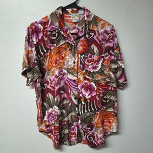 May include: A short-sleeved button-down shirt with a vibrant floral pattern in shades of purple, orange, and brown. The shirt features a collared neckline and a pocket on the chest. The tag reads "Ki Ko Mo."