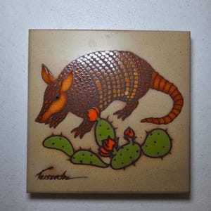 May include: Square ceramic tile with an armadillo design in orange and brown, alongside a green cactus with orange flowers. The tile has a beige background and a signature. Decorative tile, approximately 15 cm square.