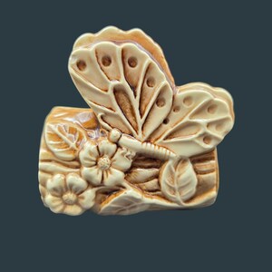 May include: A cream-colored ceramic decorative object featuring a sculpted butterfly resting on a textured log surface adorned with two small flowers and leaves. The piece has a glossy finish with brown shading in the recessed areas of the design.