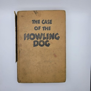 May include: A vintage book with a tan cover. The title, "THE CASE OF THE HOWLING DOG," is printed in bold black letters. The book shows signs of wear, with a slightly torn spine and some discoloration.