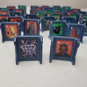 May include: A collection of blue plastic game pieces featuring character illustrations and the text "DARK SIDE". The game pieces have a rectangular card with a character image and a number. The background is blurred, showing more game pieces.
