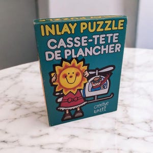 May include: A teal box for an Inlay Puzzle, with text in yellow and white. The box features a cartoon sun character in a red dress and a helicopter with a superhero. The brand is Creative Kids.
