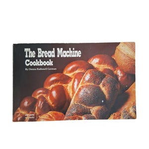 May include: A vintage cookbook titled "The Bread Machine Cookbook" by Donna Rathmell German. The cover features a dark background with various types of freshly baked bread. The bread is golden brown with poppy seeds on top.