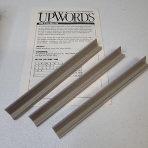 May include: A board game titled "UPWORDS" with instructions and letter distribution details. The image shows three tan-colored game board rails. The game is designed for 2 to 4 players and involves stacking letter tiles to form words.