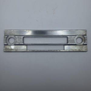 May include: A silver-colored, rectangular metal plate with a central rectangular opening and two circular openings on either end. The plate has a slightly curved shape and a textured surface with horizontal lines.