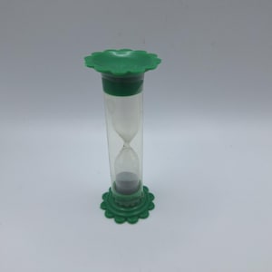 May include: A green and clear hourglass with a flower-shaped top and base. The hourglass is made of clear glass and green plastic. The sand inside is dark gray. The hourglass is a timekeeping device.