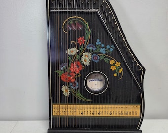 Puede incluir: Una cítara negra con un diseño floral de flores rojas, blancas, azules y amarillas. El instrumento tiene una boca circular con el texto Jubeltöne, clavijas de afinación de metal y una tira guía musical amarilla con notas musicales.