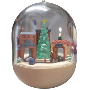 May include: A clear plastic snow globe ornament with a miniature winter village scene inside. The scene includes a green Christmas tree, a building with the word "Hallmark" on it, and a building with the year "1993" on it.