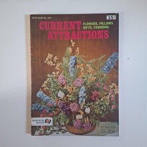 May include: A vintage book cover titled "Current Attractions" with a floral arrangement. The cover features text that reads "Flowers, Pillows, Gifts, Fashions." The book is from Star Book No. 214 and includes the text "All made with Aunt Lydia's heavy rug yarn."