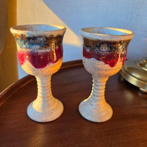 May include: Two ceramic goblets featuring a speckled cream base, a ribbed stem, and a wide bowl. The upper portion of each bowl displays a horizontal band of deep red glaze topped with a textured, mottled blue and brown drip pattern.