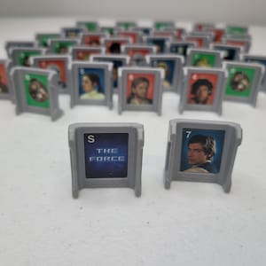 May include: A collection of gray plastic game pieces featuring images of characters and text from the Star Wars franchise. One piece displays the text "THE FORCE" and another shows a portrait of Han Solo with the number "7".