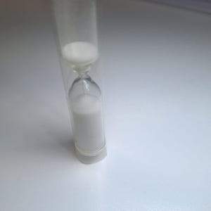 Könnte beinhalten: Eine transparente Glasuhr, gefüllt mit weißem Sand. Die Sanduhr hat eine schmale Mitte und breitere obere und untere Abschnitte. Der Sand befindet sich teilweise im oberen Abschnitt, der Rest im unteren Abschnitt. Die Sanduhr ist in einem transparenten Kunststoffhalter eingeschlossen.