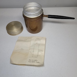 May include: A copper coffee maker with a black handle, a removable silver-toned metal strainer lid, and a separate perforated metal cap. Included is a folded paper instruction sheet labelled Douro Copperware, Made in Portugal, How to enjoy true Italian Coffee.