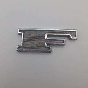 以下が含まれることがあります： 文字「F」の形をした銀色の金属エンブレム。エンブレムは、文字の形の中にテクスチャ加工された溝のある表面を持っています。背景は無地の明るい灰色です。