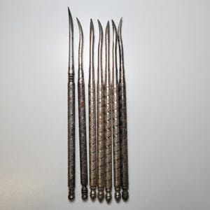 May include: A collection of antique silver tools, likely for a specific craft. The tools have slender, curved tips and ornate, textured handles. The tools are arranged vertically against a white background, showcasing their detailed craftsmanship.