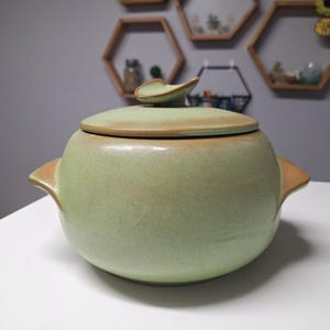 May include: A green ceramic casserole dish with a lid. The dish has a rounded shape and two handles on the sides. The lid has a leaf-shaped knob.