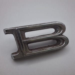 May include: A vintage, chrome-plated letter "B" emblem. The emblem is rectangular with a stylized "B" shape, featuring a double-lined design. The metal has a slightly aged appearance, with some surface wear. The emblem is likely from a car or vehicle.