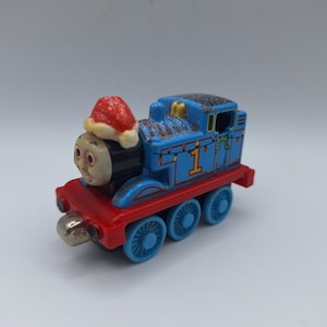 Op de afbeelding: Een blauwe speelgoedtrein met het nummer 1 aan de zijkant, een rode basis en blauwe wielen. De trein draagt een rode kerstmuts met witte rand. De trein heeft een lachend gezicht en is versierd met groene en gele lichtjes.