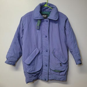 May include: A light purple winter coat with a large collar and two front pockets. The coat has a button-up closure and a green lining visible on the collar and pockets. The brand name "ZZI" is visible on the collar.