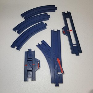 May include: A collection of blue plastic toy train track pieces, including three curved segments, one straight switch track with a red lever, and one straight track section featuring a red sliding mechanism for a toy railway system.