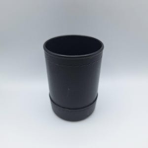 May include: A black dice cup with a textured surface. The cup is cylindrical, featuring a stitched seam near the top and a wider base. The interior is dark, set against a plain white background. The cup is approximately 10 cm tall.