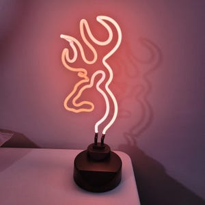 May include: A neon light sculpture in the shape of a deer head with antlers. The light is a warm orange color and sits on a black base. The sculpture casts a shadow on the wall behind it.