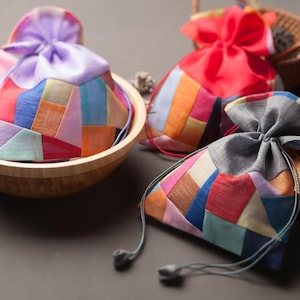 Op de afbeelding: Een verzameling kleurrijke patchwork stoffen zakjes met trekkoord sluitingen. De zakjes zijn in verschillende kleuren, waaronder roze, paars, rood en blauw. Ze zijn gerangschikt in een houten kom en op een donkere ondergrond, wat wijst op een ambachtelijk of cadeau-artikel.