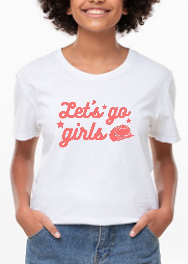 May include: White t-shirt with a pink graphic that says "Let's go girls" with a pink cowboy hat graphic.