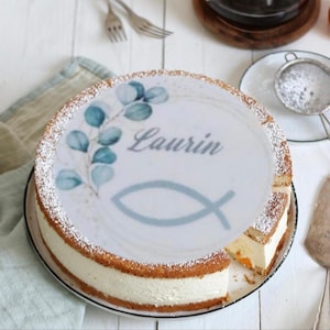 Könnte beinhalten: Ein runder Kuchen mit weißer Füllung und hellbrauner Kruste. Die Oberseite des Kuchens hat ein essbares weißes Bild mit blauen Blättern, dem Namen "Laurin" und einem blauen Fischsymbol. Der Kuchen ist mit Puderzucker bestäubt und liegt auf einem weißen Teller.