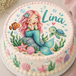 Könnte beinhalten: Ein runder Kuchen mit weißer Glasur, verziert mit einer Meerjungfrauen-Illustration, einem Clownfisch, einer Meeresschildkröte, Seesternen und Muscheln. Der Name Lina ist in türkisfarbener Schrift gedruckt. Der Rand hat weiße Sahnetupfen.