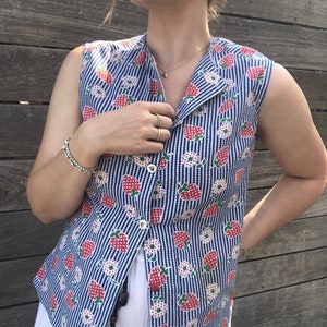May include: Sleeveless vest with a blue and white striped pattern, adorned with red strawberries and white flowers. The vest has a collared neckline and red buttons. The garment is part of a summer clothing collection.