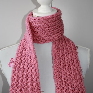May include: A pink knitted scarf, displayed on a mannequin. The scarf has a textured, open weave design. The scarf is wrapped around the mannequin's neck and hangs down. The scarf is a solid colour.