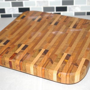 Puede incluir: Tabla de cortar cuadrada de madera con esquinas redondeadas. Está hecha de varios tonos de madera, incluyendo marrones claros y oscuros, con algunos acentos más oscuros. La veta de la madera es visible. Mide aproximadamente 30 cm x 30 cm.