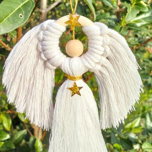 Puede incluir: Adorno de ángel de macramé hecho a mano con alas de flecos blancos y una cabeza de cuentas de madera. El ángel tiene un halo de anillo de madera y detalles de estrella dorada. Se utiliza un hilo dorado para colgar, ideal para decoración navideña.