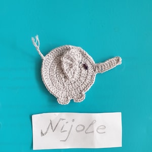 May include: A crocheted elephant in light gray yarn, featuring a trunk, ears, and small legs. The elephant has a small purple bead for an eye. A handwritten note below reads "Nijole".