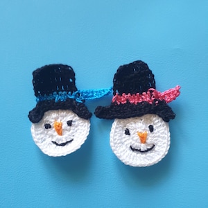 Könnte beinhalten: Zwei gehäkelte Schneemann-Gesichter mit schwarzen Zylindern, orangenen Karottennasen und lächelnden Mündern. Einer hat einen blauen Schal, der andere einen rosa Schal. Die Schneemänner sind weiß mit schwarzen Augen, vor blauem Hintergrund.