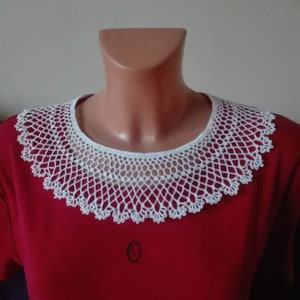 May include: White crocheted collar with a delicate pattern. The collar is worn on a red shirt.
