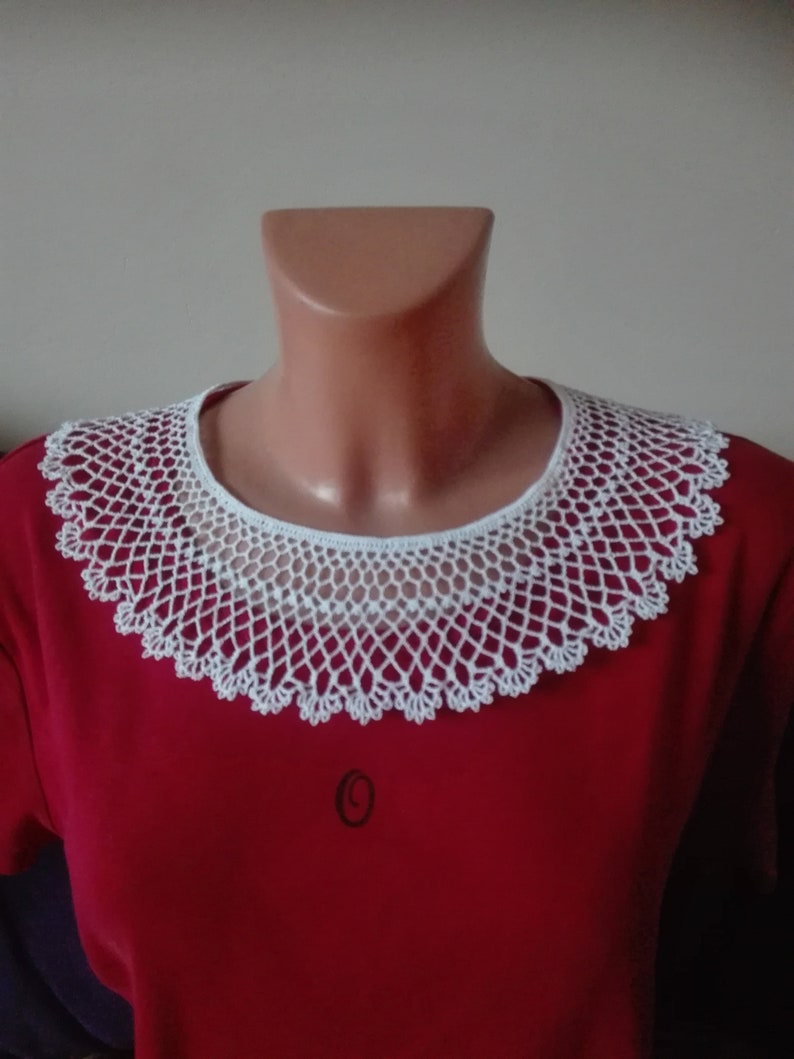 May include: White crocheted collar with a delicate pattern. The collar is worn on a red shirt.