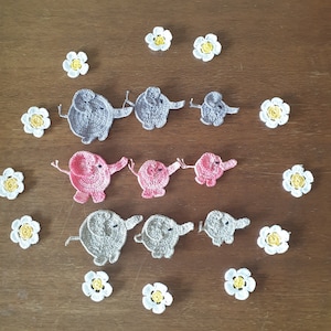 May include: A set of nine crocheted elephants in gray and pink, arranged in three rows of three. The elephants are surrounded by white crocheted flowers with yellow centers.