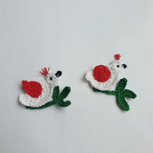 May include: Two handmade crochet bird appliques. Each bird is white with a red wing and a red crest. They have black eyes and are perched on green crocheted branches. The background is plain white.