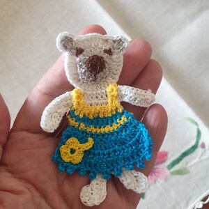 Op de afbeelding: Een gehaakte witte teddybeer die een blauwe en gele jurk draagt met een gele bloem op de voorkant.