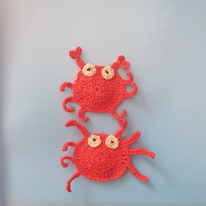 Peut inclure: Deux appliqués de crabes au crochet rouges faits à la main. Chaque crabe a un corps rond, plusieurs pattes et deux yeux avec des centres beiges. Les crabes sont affichés sur un fond bleu clair.