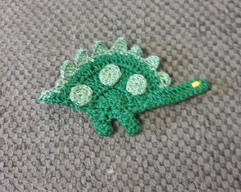 May include: A green crocheted dinosaur toy with a yellow tail. The dinosaur has a spiky back and a round body.