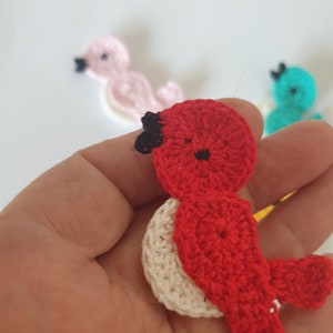 May include: A close-up of a handmade red crochet bird with a white belly and black beak. Other crochet birds in pink and teal are slightly out of focus in the background. The bird is held in a hand.